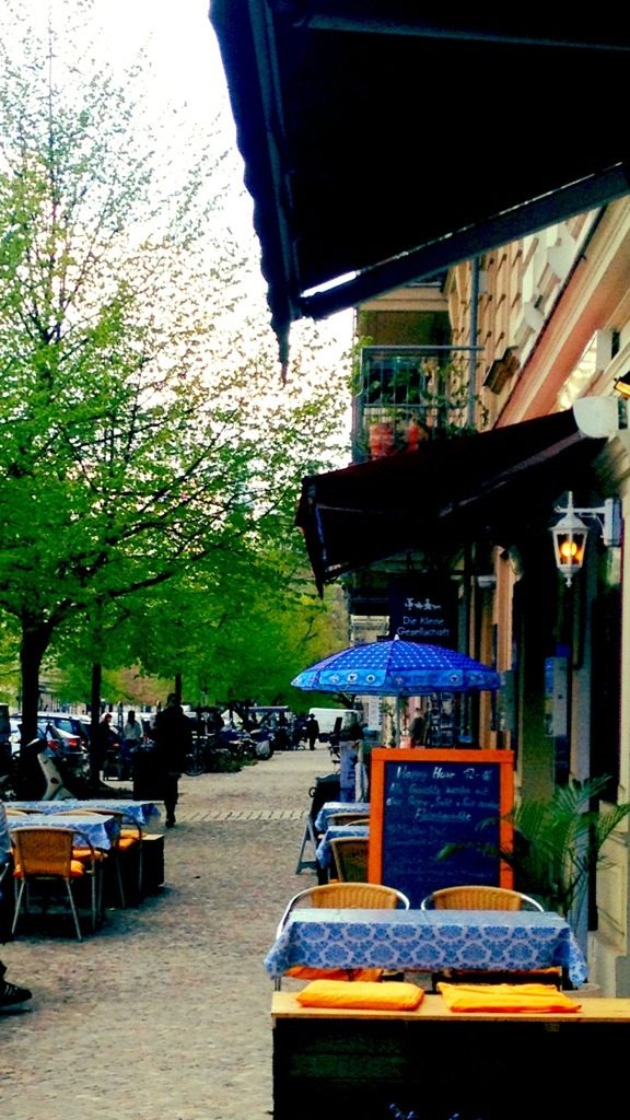 Rykestraße, Straßenansicht mit Bäumen, blauer Sonnenschirm vor der Kleinen Gesellschaft